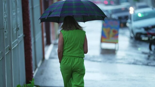 Back of woman walking in the rain holding umbrella. Person strolling during rainy day covered against bad weather