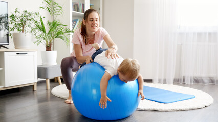 Young caring mother stretching muscles of her baby son and exercises on fitness ball