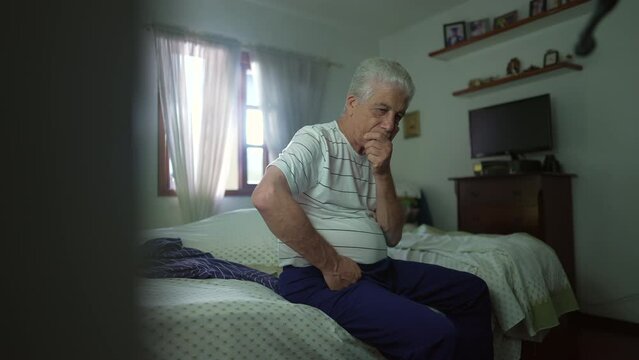 Pensive senior man seated by bedside in contemplation. Retired elderly person pondering decision with hand in chin in deep mental reflection in casual bedroom