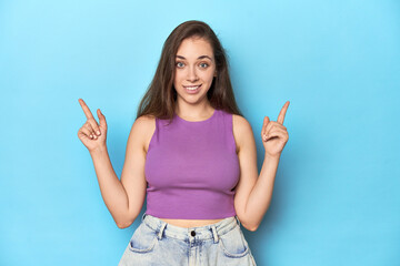 Fashionable young woman in a purple top on blue background indicates with both fore fingers up...