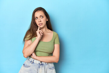 Young Caucasian woman in a green top on a blue backdrop looking sideways with doubtful and skeptical expression.