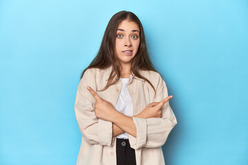 Stylish young woman in an overshirt on a blue background points sideways, is trying to choose between two options.