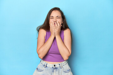 Fashionable young woman in a purple top on blue background laughing about something, covering mouth with hands.