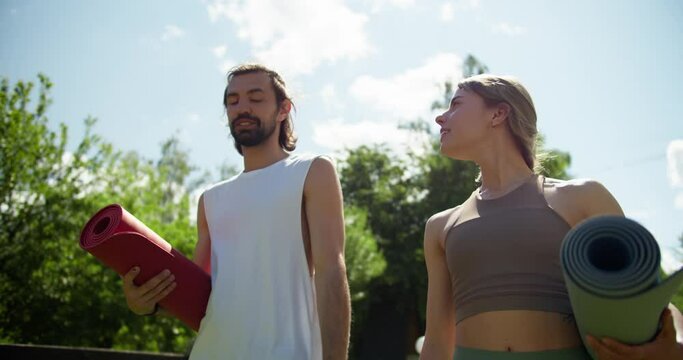 A blonde girl and a brunette guy in a summer sports uniform are walking through the park and getting ready for yoga in nature. The guy and the girl are specially mats for yoga and communicate in the