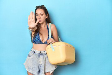 Woman in bikini with a portable beach cooler, blue studio standing with outstretched hand showing...