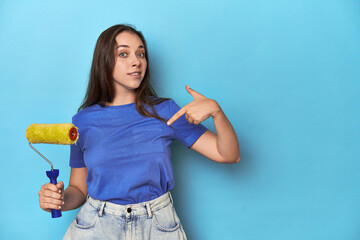 Young woman with yellow paint roller on a blue background person pointing by hand to a shirt copy space, proud and confident