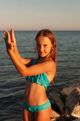 A beautiful girl in a blue swimsuit at sunset on the seashore looks at the camera and makes a figure with her hands