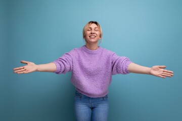 smiling cute blond young woman in a lilac sweater shows reaching out to hug on a bright background with copy space