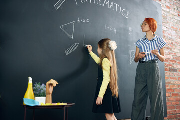Little smart girl standing by chalkboard with teacher, writing and solving math tasks. Geometry...