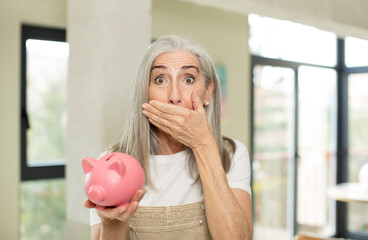 pretty senior woman covering mouth with a hand and shocked or surprised expression. with a piggy...