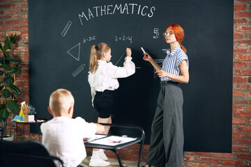 Young woman, teacher of math standing by blackboard with little girl student and solving geometry...