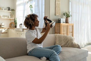 Millennial African American girl with curly hair sitting on couch testing new VR headset, using...
