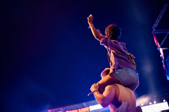 Rear view of man carrying his girlfriend on his shoulders during open air music concert at night.