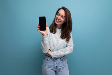 well-groomed young brunette female adult in a striped shirt and jeans shows a phone mockup isolated on a plain background