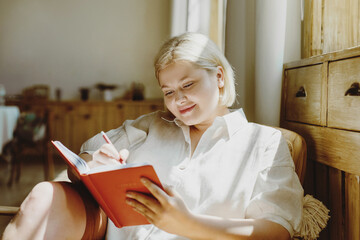 Cheerful young woman making notes in paper notebook sitting on cozy chair at home.