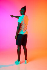 Fototapeta na wymiar Full length image of backview african man in glasses holding his hand up and pointing something over orange-pink background in neon.