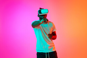 Young african american man play video games in VR-glasses showing his fist to camera over pink-orange background in neon.