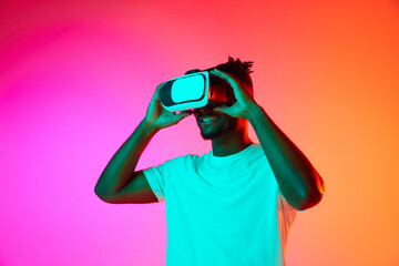 Half length image of african man smiling while playing video games in VR-glasses over pink-orange background in neon.