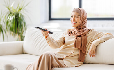 Cheerful muslim woman in hijab holding tv remote and watching series  at home.