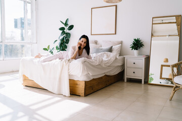 Obraz na płótnie Canvas Positive woman talking on smartphone in bedroom after awakening