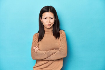 Young Asian woman in brown turtleneck, frowning face in displeasure, keeps arms folded.