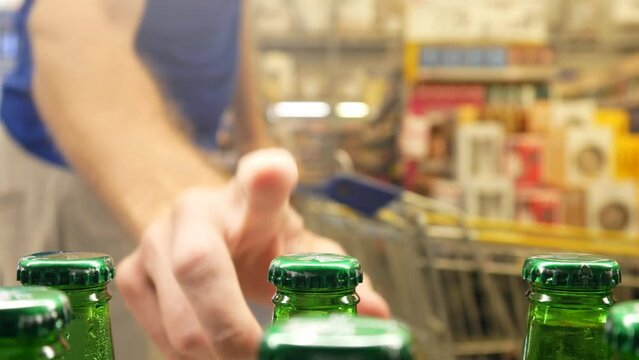 Close-up of many green glass bottles of lager beer in alcohol department and a man with a shopping trolley takes one
