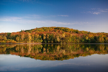 Autumn landscape. A lake with a smooth mirror surface of water and a forest on the shore in autumn colors. Deciduous forest in orange tones on the shore of the purest lake.