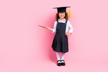 Full size photo of clever positive schoolkid wear mortarboard hat hand hold pointed indicate empty space isolated on pink color background
