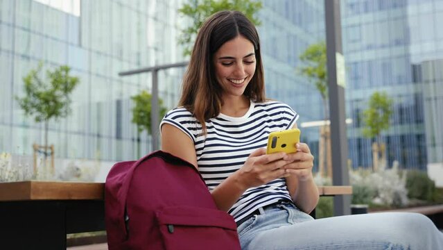 Happy hispanic student girl using yellow cell phone outdoors at University campus. Smiling young woman having fun with online app 