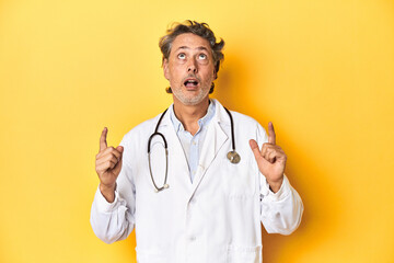 Middle-aged doctor standing against a yellow backdrop pointing upside with opened mouth.