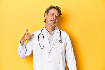 Middle-aged doctor standing against a yellow backdrop person pointing by hand to a shirt copy space, proud and confident