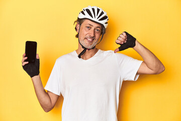 Cyclist man showing phone on yellow backdrop feels proud and self confident, example to follow.
