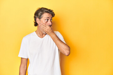 Middle-aged man posing on a yellow backdrop thoughtful looking to a copy space covering mouth with...