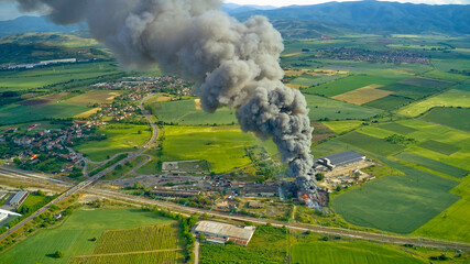 Fototapeta premium A black column of smoke and a fire in a warehouse, against the backdrop of the city. Photo from above from a drone.