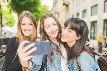 Three young beautiful caucasian young women millennials, taking selfie with smart phone hand hold outdoor in the city, smiling - social network, technology, friendship concept