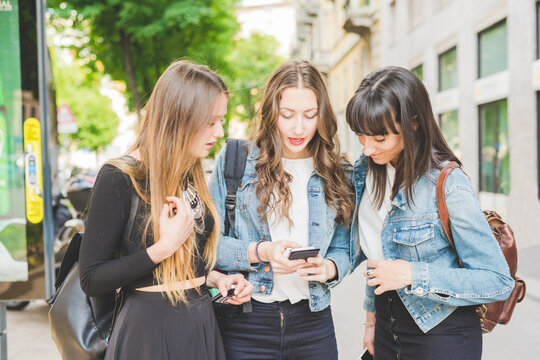 Three young beautiful caucasian women millennials outdoor in the city using smart phone hand hold, looking down and tapping the screen - technology, social network, communication concept