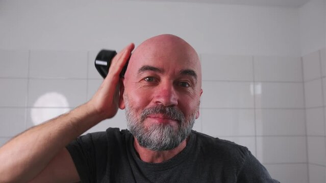 Senior man shaves his bald head with an electric razor. A man who has lost his hair takes care of his hair and scalp.