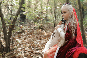 A child girl sits on a stump in the forest in a medieval dress in the form of a dead princess with makeup on her face and hugs a skeleton doll