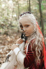 A child girl sits on a stump in the forest in a medieval dress in the image of a dead princess with makeup on her face, hugs a skeleton doll and looks into the camera.
