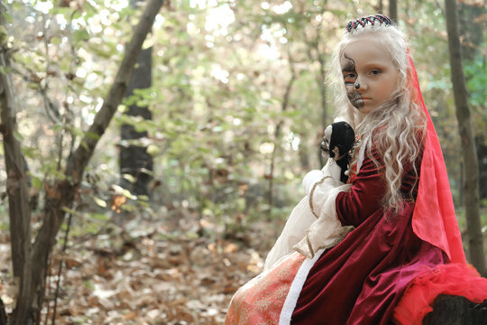 A child girl sits on a stump in the forest in a medieval dress in the image of a dead princess with makeup on her face, hugs a skeleton doll and looks into the camera.