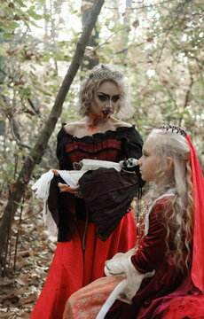 A woman in the form of a vampire or a sorceress holds a doll in the form of a skeleton in her arms and looks at a princess girl in fairy-tale makeup and medieval dress. Image for Halloween. 