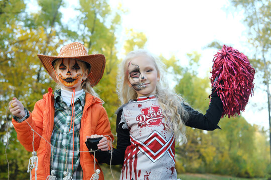 A girl in a cheerleader costume with half-face skeleton makeup and a girl with pumpkin makeup in a hat pose and look at the camera