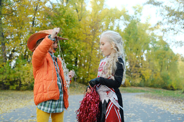 Girl child in an orange jacket and a cowboy hat shows her friend in a cheerleader costume her...