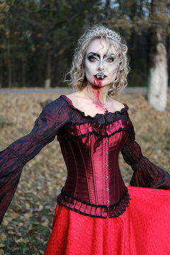 A woman in the image of a vampire or a witch with white make-up and in a medieval dress poses in the forest in front of the camera
