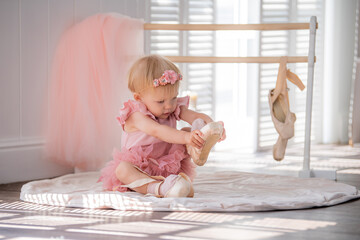 A cute little ballerina in a pink ballet costume sits near the barre in the room and tries to put on her pointe shoes. Kid and ballet dance. Copyspace.