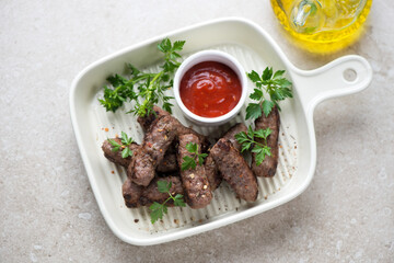 Beige serving tray with bbq serbian cevapi or cevapcici, fresh parsley and dip, horizontal shot on a beige stone background, high angle view