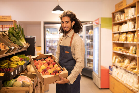 Smiling ethnic male worker showing box with ripe peaches in store