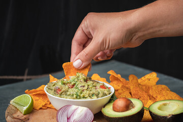 Woman hand dip nachos into tasty guacamole sauce on the wooden background. Tortilla chips next to guacamole ingredients: avocado, cilantro, onion, hot pepper and lime. Traditional Mexican food