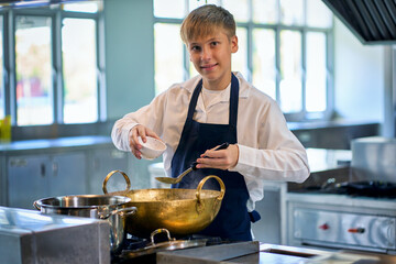 Single young boy in chef uniform try to cooking food with brass pot in kitchen for make a soup menu.