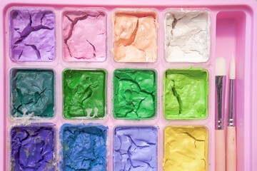 Dried colorful gouache in pink painting palette box with two pieces of white paint brush. Cracked water color and small brush.